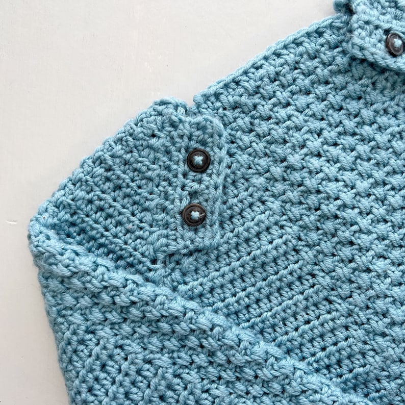 May include: A close-up of a light blue crocheted sweater with two black buttons on the shoulder.