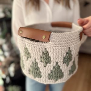 May include: A beige crochet basket with a repeating green tree pattern. The basket has brown leather handles secured with brass rivets. The basket is held by a person.