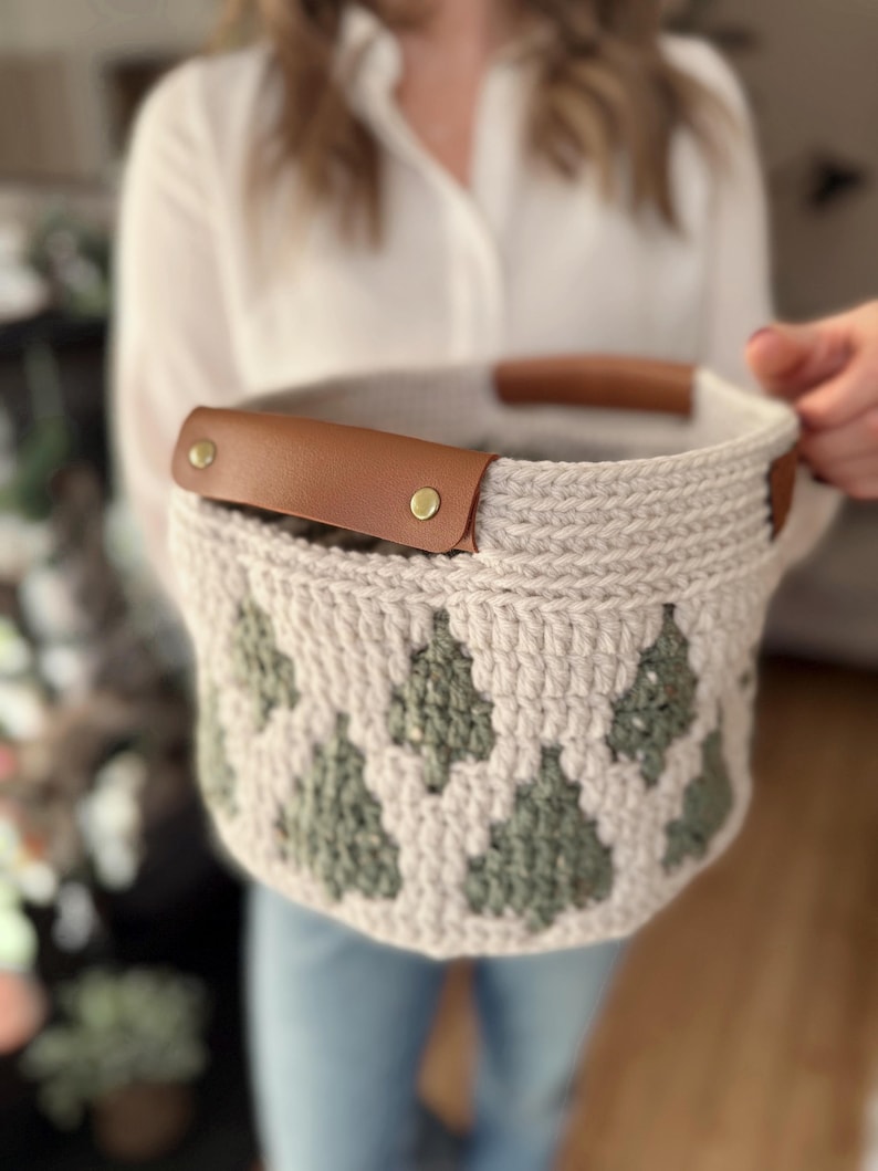 May include: A beige crochet basket with a repeating green tree pattern. The basket has brown leather handles secured with brass rivets. The basket is held by a person.
