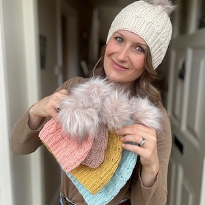 Könnte beinhalten: Eine Sammlung gestrickter Wintermützen in verschiedenen Farben, darunter Rosa, Gelb und Blau, jeweils mit einem Kunstfell-Bommel. Die Mützen werden in verschiedenen Stilen präsentiert, mit einer cremefarbenen Zopfstrickmütze mit Bommel oben.