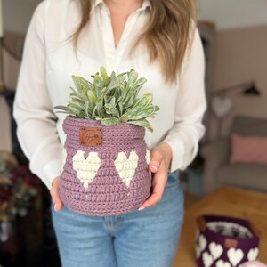 Op de afbeelding: Een gehaakte lavendelkleurige mand met witte hartpatronen, met een groene plant. De mand heeft een klein bruin label. Extra manden met hartpatronen zijn zichtbaar op de achtergrond.