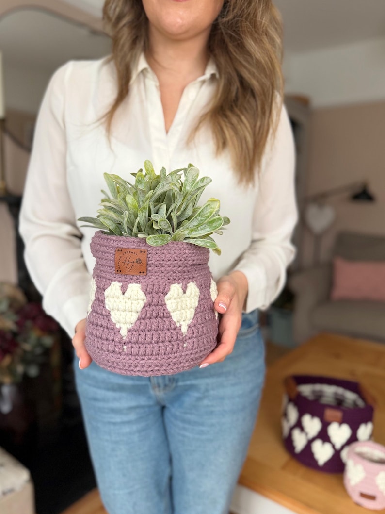 Op de afbeelding: Een gehaakte lavendelkleurige mand met witte hartpatronen, met een groene plant. De mand heeft een klein bruin label. Extra manden met hartpatronen zijn zichtbaar op de achtergrond.