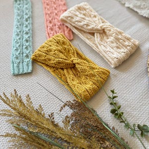 May include: A collection of handmade knitted headbands in various colors, including light blue, pink, cream, and mustard yellow. The headbands feature intricate cable knit and twisted knot designs. The image also includes dried ornamental grasses.