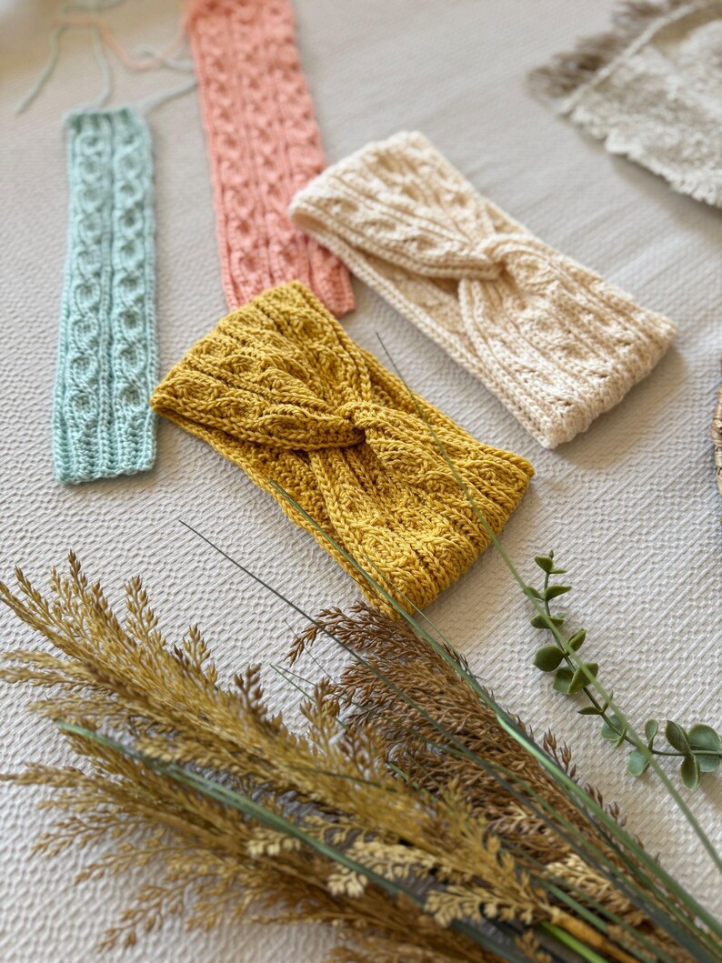 May include: A collection of handmade knitted headbands in various colors, including light blue, pink, cream, and mustard yellow. The headbands feature intricate cable knit and twisted knot designs. The image also includes dried ornamental grasses.