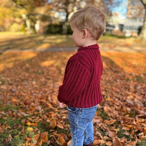 K&ouml;nnte beinhalten: Ein kleines Kind in einem weinroten Strickpullover und blauer Jeans steht in einem Haufen Herbstlaub.