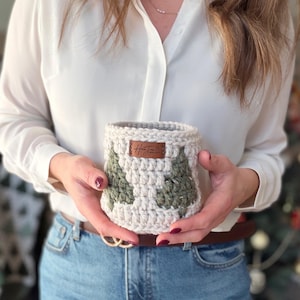 May include: A cream and green crochet basket with a tree design. The basket has a brown leather tag with text. The person holding the basket is wearing a white shirt and blue jeans. The basket is handmade.