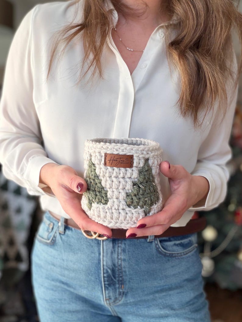 May include: A cream and green crochet basket with a tree design. The basket has a brown leather tag with text. The person holding the basket is wearing a white shirt and blue jeans. The basket is handmade.