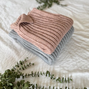May include: Two folded jumpers, one in dusty rose and one grey, are stacked on a white textured surface. The jumpers have a ribbed knit design. Greenery is placed in the foreground.