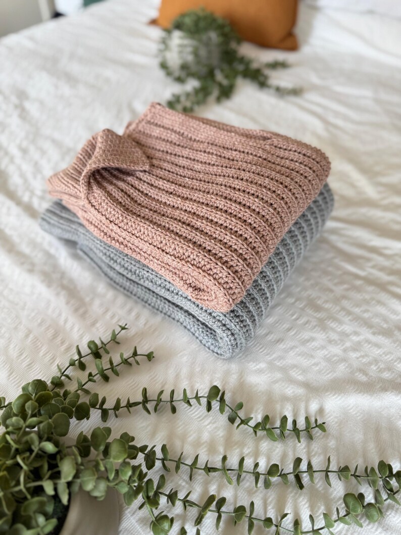 May include: Two folded jumpers, one in dusty rose and one grey, are stacked on a white textured surface. The jumpers have a ribbed knit design. Greenery is placed in the foreground.