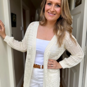 May include: An open-front cream-colored cardigan featuring a crochet knit pattern with long sleeves and ribbed cuffs. The garment is worn over a plain white top paired with white trousers and a brown leather belt with a gold-toned buckle.