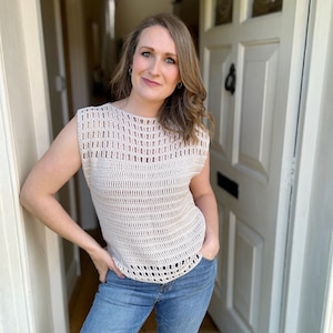 May include: A woman wearing a crocheted sleeveless top with a light brown color and a loose fit. The top has a delicate openwork pattern. She is wearing blue jeans.