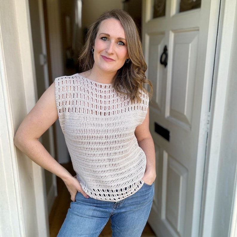 May include: A woman wearing a crocheted sleeveless top with a light brown color and a loose fit. The top has a delicate openwork pattern. She is wearing blue jeans.