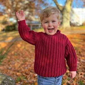 K&ouml;nnte beinhalten: Ein kleines Kind tr&auml;gt einen roten Strickpullover und eine blaue Jeans, l&auml;chelt und winkt in einem Park mit heruntergefallenen Herbstbl&auml;ttern.