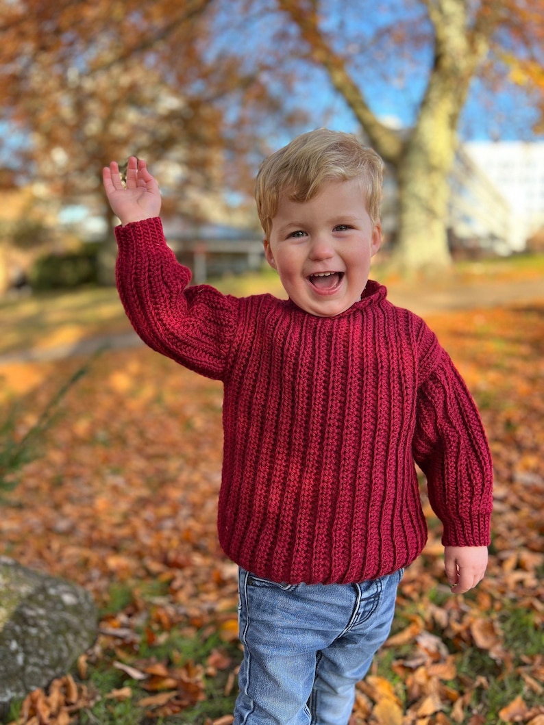 K&ouml;nnte beinhalten: Ein kleines Kind tr&auml;gt einen roten Strickpullover und eine blaue Jeans, l&auml;chelt und winkt in einem Park mit heruntergefallenen Herbstbl&auml;ttern.