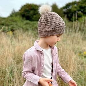 May include: A brown knit beanie with a fluffy pom-pom. The hat is worn by a person wearing a pink button-down shirt and a white t-shirt. The person is holding a woven basket while standing in a field of tall grass.