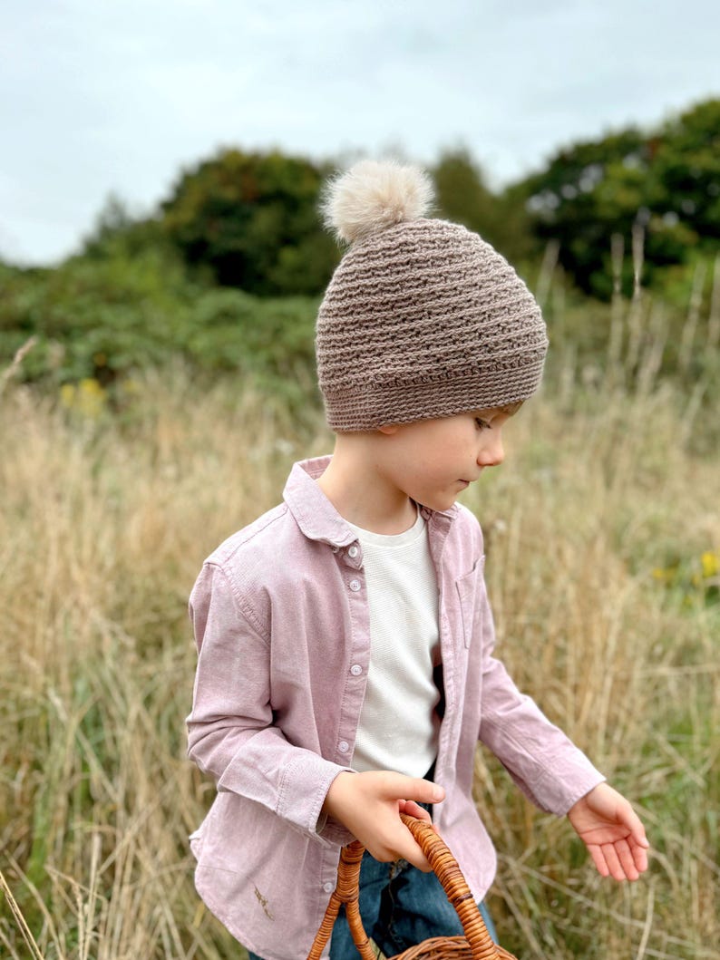May include: A brown knit beanie with a fluffy pom-pom. The hat is worn by a person wearing a pink button-down shirt and a white t-shirt. The person is holding a woven basket while standing in a field of tall grass.