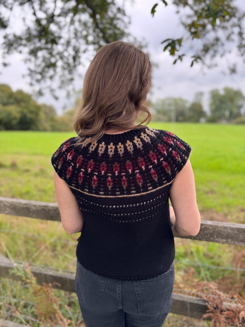 Pu&ograve; includere: Un top nero all'uncinetto con uno sprone decorativo con un motivo bordeaux, beige e nero. Il top &egrave; senza maniche e ha uno scollo tondo. La persona &egrave; in piedi fuori vicino a una staccionata di legno.