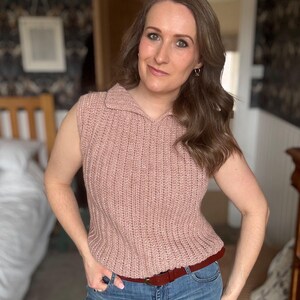 May include: A sleeveless, pale pink knitted top with a collared neckline. The top is worn with blue jeans and a brown belt. The outfit is casual and appears to be handmade.