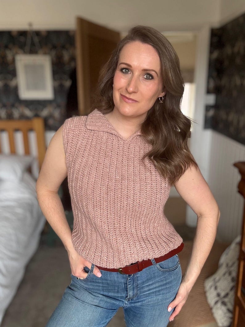 May include: A sleeveless, pale pink knitted top with a collared neckline. The top is worn with blue jeans and a brown belt. The outfit is casual and appears to be handmade.