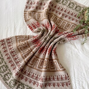 May include: A beige, brown, red, and green crochet blanket with detailed geometric patterns. The blanket is folded, displaying its design. The blanket is made with a variety of stitches, creating a textured appearance. The blanket is draped over a white surface.