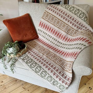May include: A cream-coloured, patterned throw blanket draped over a grey armchair. The blanket features geometric designs in red, brown, and green. A rust-coloured velvet cushion rests on the chair, with a small plant and tray visible.