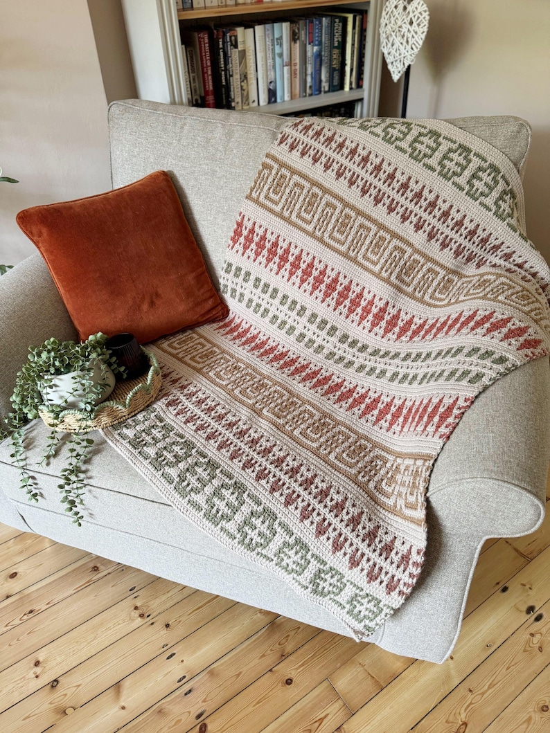 May include: A cream-coloured, patterned throw blanket draped over a grey armchair. The blanket features geometric designs in red, brown, and green. A rust-coloured velvet cushion rests on the chair, with a small plant and tray visible.