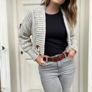 Pu&ograve; includere: Una donna indossa un cardigan corto color crema con una maglietta nera e jeans grigi. Indossa una cintura in pelle marrone con una fibbia dorata.