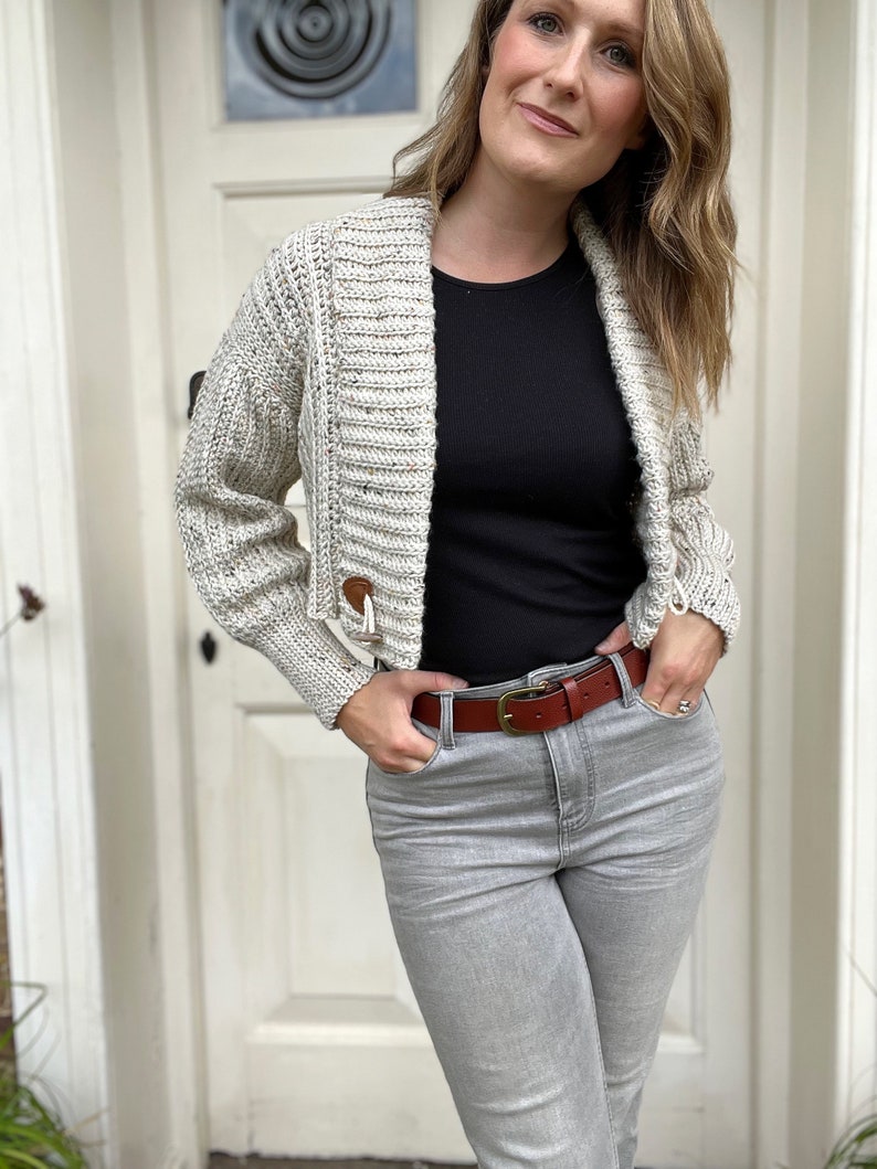 Pu&ograve; includere: Una donna indossa un cardigan corto color crema con una maglietta nera e jeans grigi. Indossa una cintura in pelle marrone con una fibbia dorata.