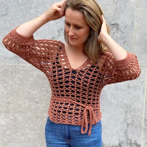 Puede incluir: Una blusa de crochet de color terracota con cuello en V y mangas largas. La blusa tiene un lazo en la cintura y se lleva sobre un top negro de tirantes.