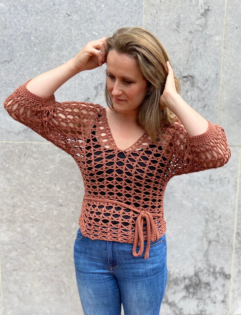 Puede incluir: Una blusa de crochet de color terracota con cuello en V y mangas largas. La blusa tiene un lazo en la cintura y se lleva sobre un top negro de tirantes.