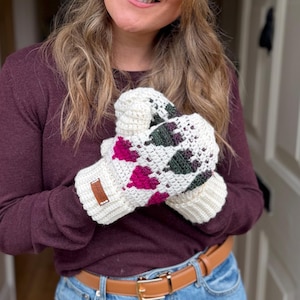 May include: Cream-colored crochet mittens with a geometric pattern of pink and green diamonds. The mittens have a ribbed cuff and a small brown tag. The mittens are held in front of a person wearing a burgundy sweater and blue jeans.