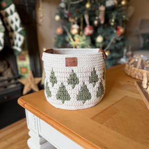 May include: A cream-coloured, crocheted basket featuring a pattern of green Christmas trees. The basket has brown leather handles and a small leather tag with initials. It sits on a wooden table, with a Christmas tree visible in the background.