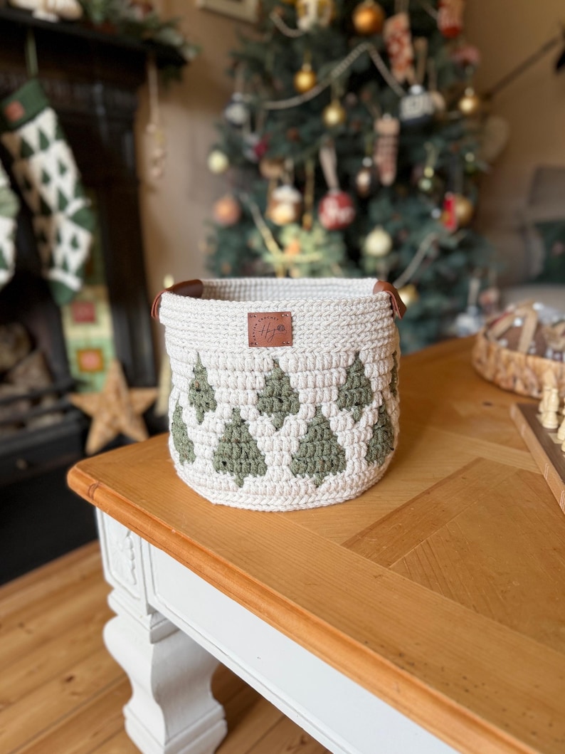May include: A cream-coloured, crocheted basket featuring a pattern of green Christmas trees. The basket has brown leather handles and a small leather tag with initials. It sits on a wooden table, with a Christmas tree visible in the background.