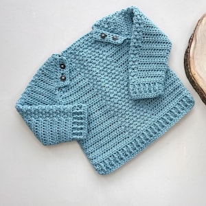 May include: A light blue crocheted sweater with a button closure at the neck. The sweater has a textured stitch pattern and is made with a soft yarn.