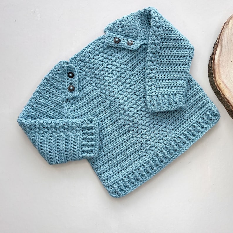 May include: A light blue crocheted sweater with a button closure at the neck. The sweater has a textured stitch pattern and is made with a soft yarn.