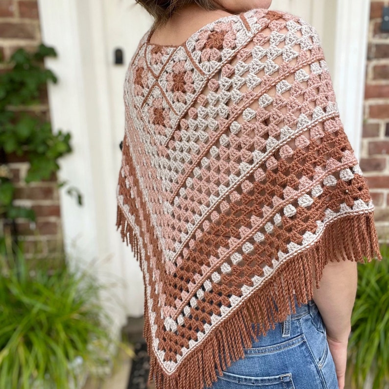 Puede incluir: Un poncho de ganchillo con un patr&oacute;n geom&eacute;trico en tonos marrones, rosas y blancos. El poncho tiene un borde de flecos.