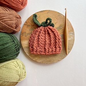 May include: A crocheted pumpkin hat in orange with a green stem and leaves. The hat is on a wooden plate with yarn balls in various shades of green, brown, and orange.