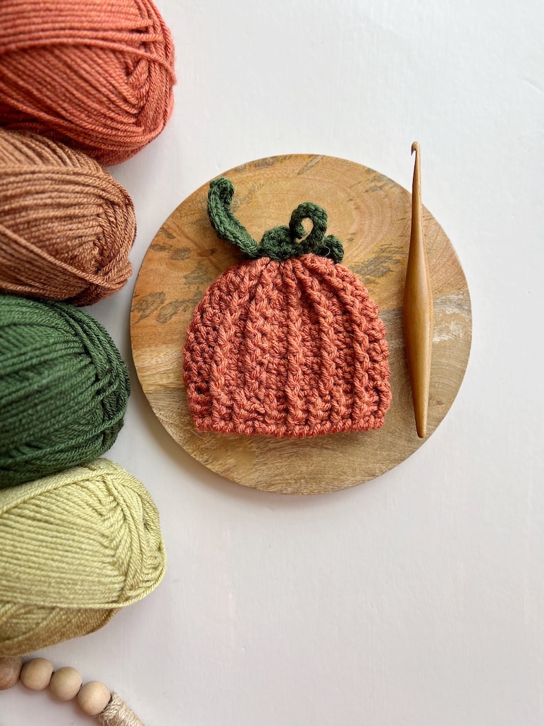May include: A crocheted pumpkin hat in orange with a green stem and leaves. The hat is on a wooden plate with yarn balls in various shades of green, brown, and orange.