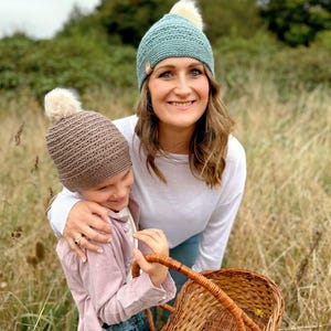 May include: Two crocheted beanies, one teal and one brown, each with a fluffy white pom-pom. The teal beanie is worn by a person, and the brown beanie is held by another person. A woven basket is in the foreground.