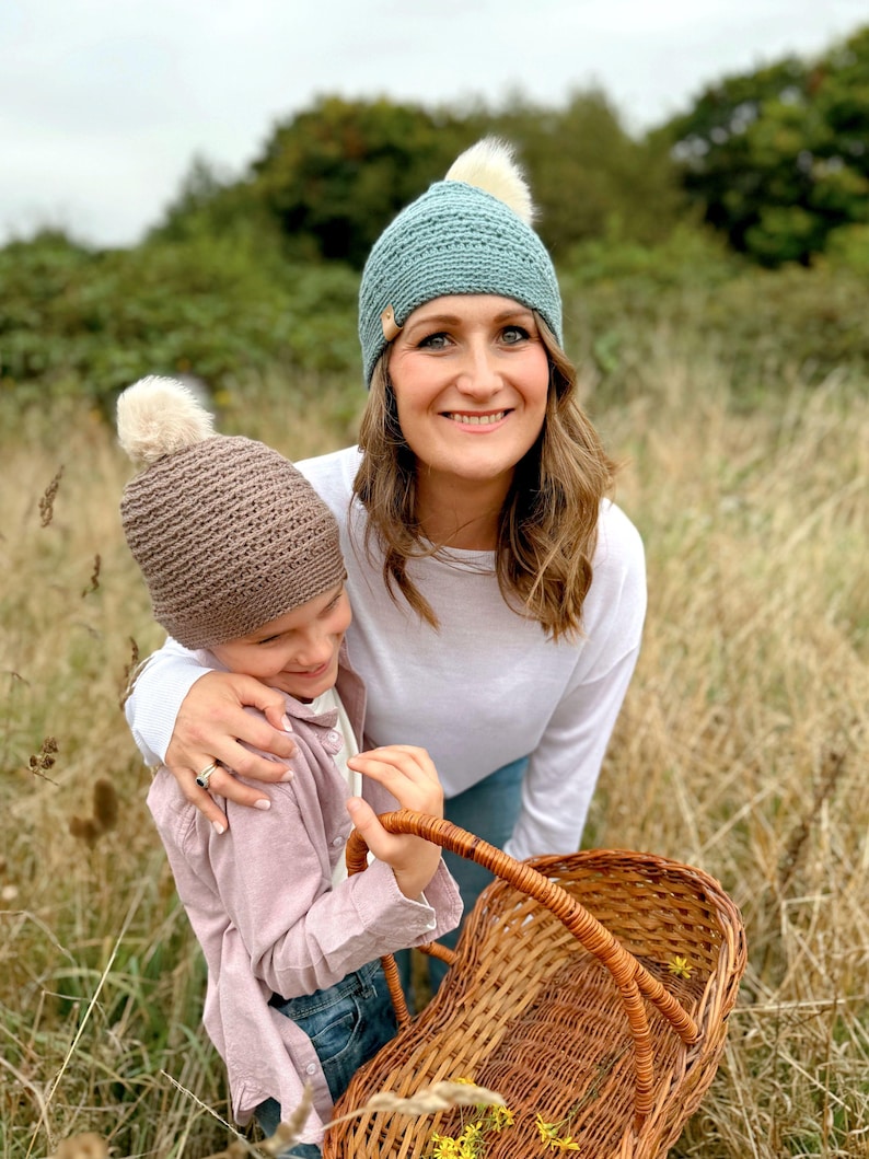 May include: Two crocheted beanies, one teal and one brown, each with a fluffy white pom-pom. The teal beanie is worn by a person, and the brown beanie is held by another person. A woven basket is in the foreground.