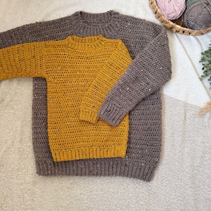Puede incluir: Dos jers&eacute;is de crochet, uno de color amarillo mostaza y otro de color marr&oacute;n oscuro, est&aacute;n apilados uno encima del otro. Los jers&eacute;is est&aacute;n hechos con un hilo grueso y tienen un dise&ntilde;o simple y cl&aacute;sico.