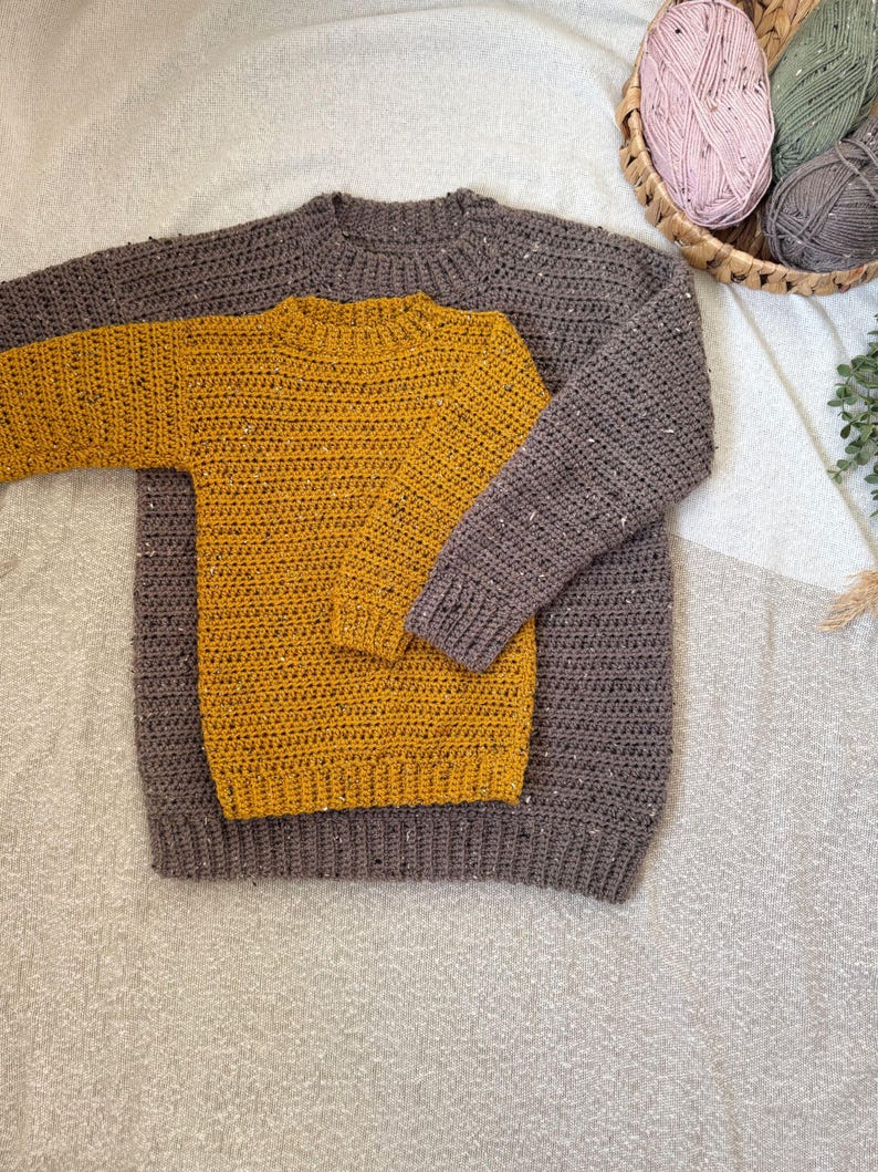 Puede incluir: Dos jers&eacute;is de crochet, uno de color amarillo mostaza y otro de color marr&oacute;n oscuro, est&aacute;n apilados uno encima del otro. Los jers&eacute;is est&aacute;n hechos con un hilo grueso y tienen un dise&ntilde;o simple y cl&aacute;sico.