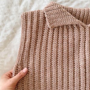 May include: Close-up of a handmade, light brown crochet top. The ribbed texture is visible, with a folded collar. The garment is displayed on a white surface. The crochet work appears to be tightly knit.