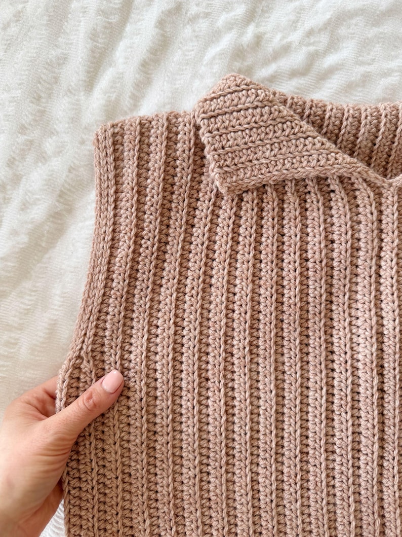 May include: Close-up of a handmade, light brown crochet top. The ribbed texture is visible, with a folded collar. The garment is displayed on a white surface. The crochet work appears to be tightly knit.
