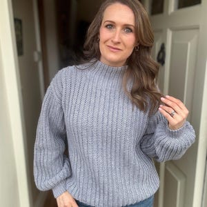 May include: A pale blue ribbed knit sweater with a high neckline and long sleeves. The sweater is worn with blue jeans. The woman is wearing a ring and earrings.