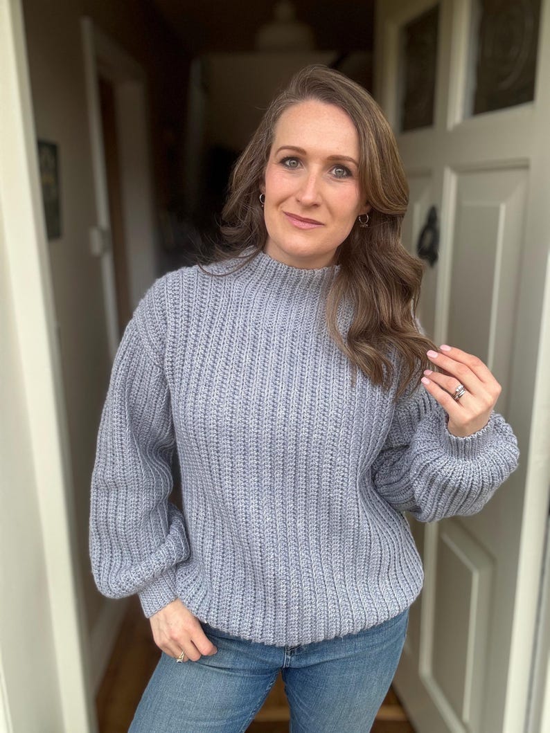 May include: A pale blue ribbed knit sweater with a high neckline and long sleeves. The sweater is worn with blue jeans. The woman is wearing a ring and earrings.