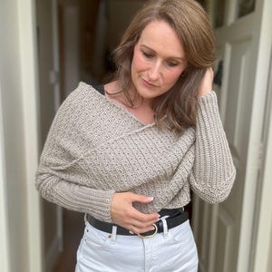 May include: A beige crocheted sweater with a draped neckline and long sleeves. The sweater is worn over white jeans and a black belt.