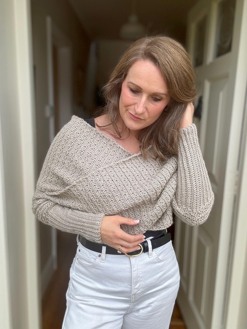 May include: A beige crocheted sweater with a draped neckline and long sleeves. The sweater is worn over white jeans and a black belt.