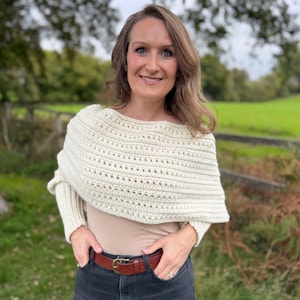May include: Cream-colored crochet shawl with a textured design. The shawl is worn over a beige top and paired with a brown leather belt and dark gray jeans. The outfit is completed with a ring.