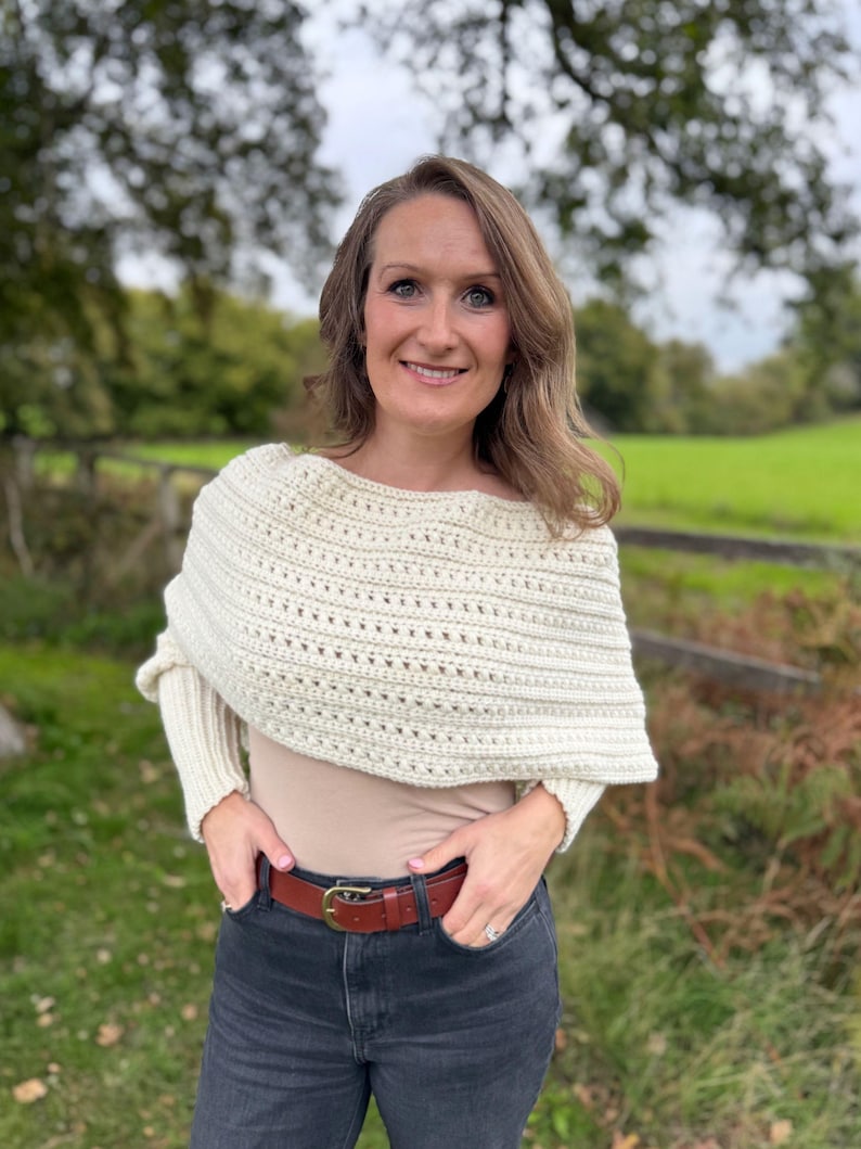 May include: Cream-colored crochet shawl with a textured design. The shawl is worn over a beige top and paired with a brown leather belt and dark gray jeans. The outfit is completed with a ring.